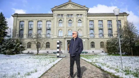 A man stands outside a building with snow on the grass