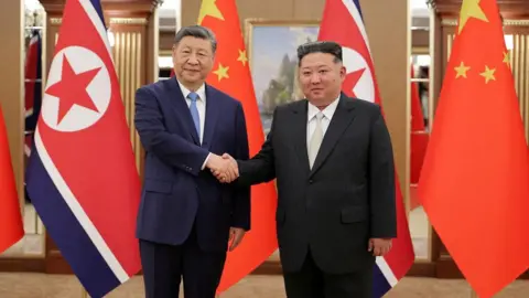 KCNA Xi Jinping and Kim Jong Un shaking hands. Both are wearing suits. Behind them is a row of Chinese and North Korean flags.
