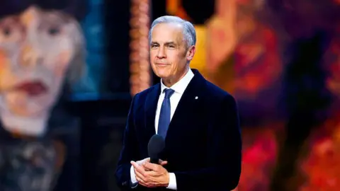 Getty Images Prime Minister Mark Carney pictured on the stage of the Juno awards in Canada. He is smiling and holding a microphone in his hand. Behind him is a blurred image of Canadian singer Joni Mitchell. He is wearing a suit and a dark blue tie.