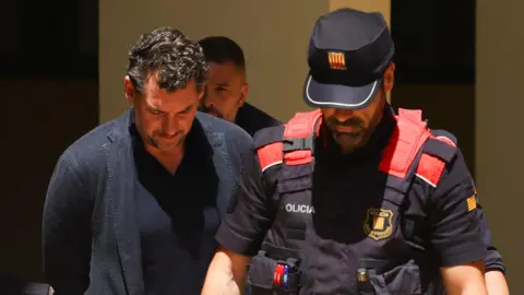 Reuters Jonathan Andic wears a blue jacket and blue top while he walks with his hands behind his back and his head down. He stands next to a policeman who is wearing a black and red uniform and cap.