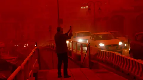 NIKOS CHALKIADAKIS/EPA/Shutterstock A man takes a picture while cars drive with their headlights on in red air.