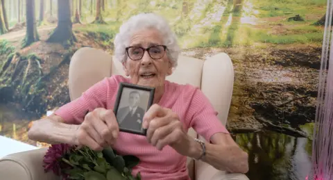 An elderly woman with white hair and glasses holding an old photograph up to the camera and wearing a pink top