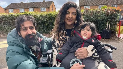 A man and woman hold a young child. The man has short, dark hair and a beard and is wearing a greyish-green padded jacket. The woman has long, dark wavy hair and is wearing a leopard-print scarf. The young child has dark hair and is wearing a dark grey padded zip-up onesie.
