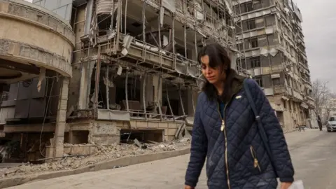 Reuters A woman walks next to bombed-out buildings on a street in Tehran (02/03/26) 