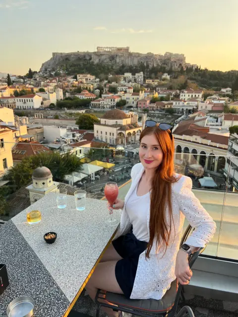 Amber Robertson Amber, a young woman with red hair, sitting at an outdoor dining table in Athens. The Acropolis is visible in the background.