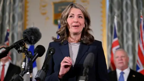 Reuters An image of Alberta Premier Danielle Smith standing and speaking at a podium. She is wearing a dark navy blue blazer with a tan top underneath and a silver necklace. She has shoulder-length brown, wavy hair.