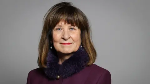 UK Parliament Baroness Helena Kennedy KC in a purple jacket with a fur collar against a neutral grey backdrop