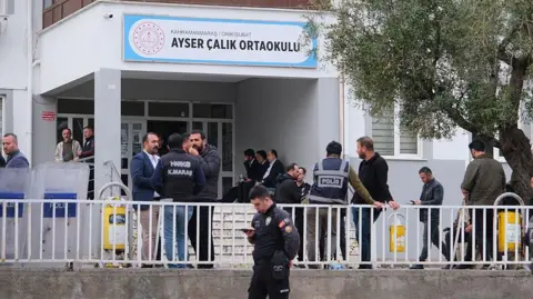 Getty Images About two dozen men stand outside a school building, which has a sign reading 'Ayser Calik Ortaokulu', wearing a mix of plain civilian clothes and police uniforms. 