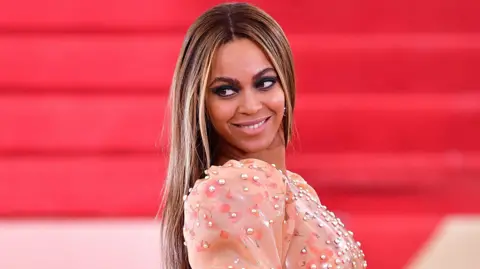 Getty Images Beyoncé Knowles wearing an embellished peach dress and with long blonde-brown hair, standing side-on and smiling off camera against a red backdrop