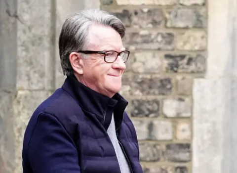 PA Peter Mandelson in grey top, navy jacket and glasses smiles as he is led away by police following his arrest earlier this week