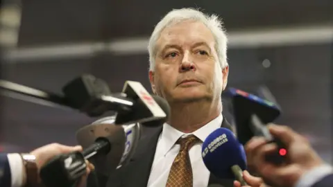 Getty Images An image showing Michael Rousseau surrounded by media microphones at a scrum in Montreal, pictured in 2021. He has short white hair, and is wearing a suit and copper patterned tie.