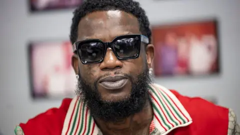 Getty Images A close-up of Gucci Mane. He wears sunglasses and a red shirt.