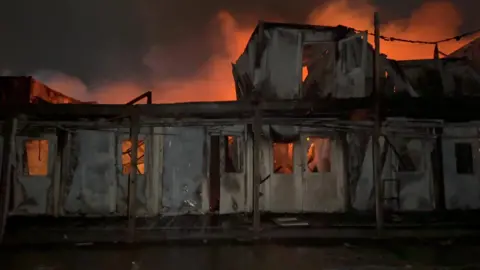 A building on fire at night. It has been badly damaged, with flames and smoke glowing orange behind the shell of the building.