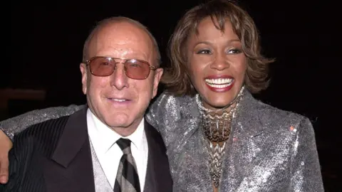 Getty Images Clive Davis in a black suit and Whitney Housten in a silver jacket with sequins smiling at the camera 