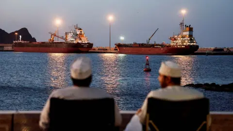 Reuters The backs of two men wearing white can be seen looking out to sea, where the Galaxy Globe bulk carrier and the Luojiashan tanker sit anchored as Iran vows to close the Strait of Hormuz, amid the U.S.-Israeli conflict with Iran, in Muscat, Oman, March 9, 2026. REUTERS/Benoit Tessier/File Photo