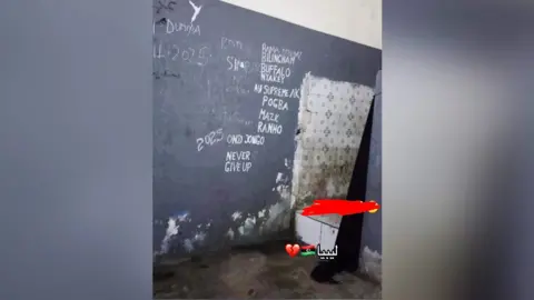 Instagram A small, worn room with damp, peeling walls and a stained floor. On the dark-painted wall, several names and words are handwritten in white, including “2025,” “Never Give Up,” and a list of names arranged vertically. In the corner, a section of tiled wall is partially visible and discolored. The lower half of a person wearing dark clothing stands near the corner, with part of the body obscured by a red mark. A small graphic with a broken heart emoji, a flag, and Arabic text appears near the bottom of the image.