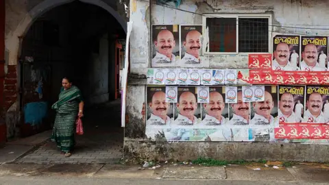 NurPhoto via Getty Images A woman walks out of a residential colony as election campaign posters of United Democratic Front (UDF) candidate Mohammed Shiyaz and Left Democratic Front (LDF) candidate K J Maxy are displayed along a roadside ahead of the Assembly election in Kochi, India, on April 4, 2026.