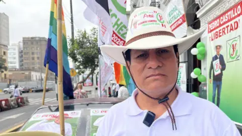 Raúl is wearing a white polo shirt and a Sánchez hat. Campaign flags can be seen behind him in the street