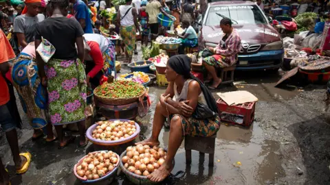 SAIDU BAH/AFP via Getty Images A busy street market scene in Freetown. There are large bowls of onions, chilli peppers and other vegetables. Women selling the produce sit on stools. One woman has her feet on the edge of a bowl of onions. Shoppers gather round to look at the items on sale. 