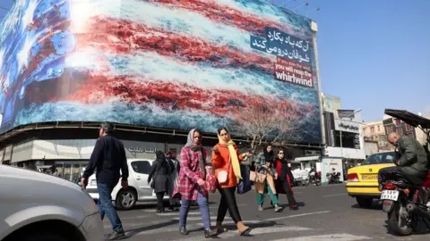 EPA Iranians walk past a billboard warning the US not to attack Iran at a square in central Tehran, Iran (25 February 2026)
