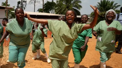 BBC Health workers wearing green scrubs and white wellie boots dance and sing on a dusty ground