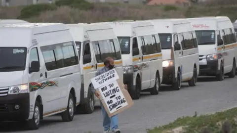 Geach/Gallo Images via Getty Images) A taxi driver stands in front of parked taxi vans in Cape Town, holding a placard that reads “No to looting, no to shooting”