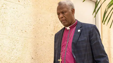 AFP via Getty Images Laurent Mbanda, wearing a purple shirt and blue jacket with a chain around his neck with a golden cross pendant