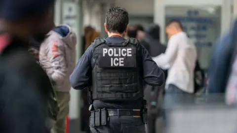 Getty Images An ICE agent walks away from the camera. He is wearing a blue-black uniform and has brown hair. The lens is in soft focus around him. 