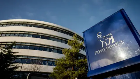 Getty Images An image showing the Novo Nordisk headquarters in Denmark. It features a circular, multi-level building with windows, trees in the front and the Novo Nordisk logo in white on a large navy blue sign. 