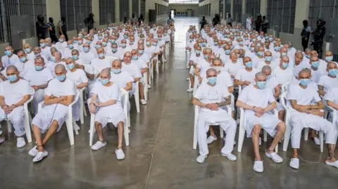 Reuters A group of hundreds of men with shaved heads, wearing white t-shirts and shorts or trousers and blue surgical masks, are seen sitting on white plastic chairs in an open space at a confinement centre in El Salvador