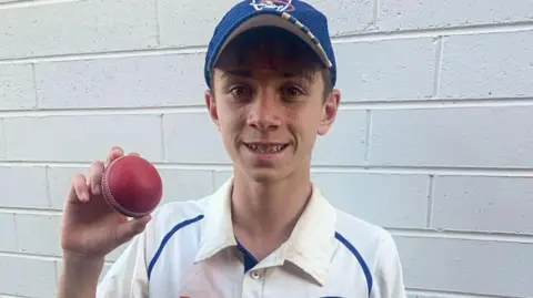 Supplied A boy wearing a blue cap and a dirty white polo poses with a cricket ball in his hand