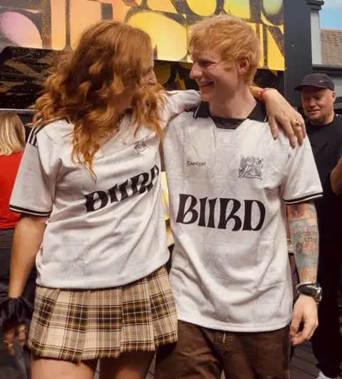 BIIRD Lisa Canny and Ed Sheeran stand close together outdoors with their arms around each other, both wearing matching white shirts with “BIIRD” printed on the front. Lisa wears a plaid skirt, while Ed has visible tattoos on one arm and is wearing brown trousers. A colorful sign or mural appears in the background, along with a few other people nearby.