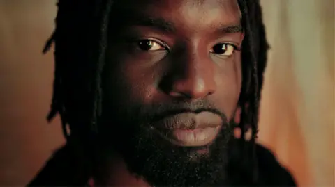 Meji Alabi A close-up of Meji Alabi, with beard and dreads, looking into the camera