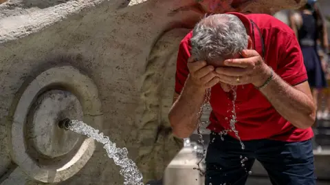 Getty Images A man cools himself at a water fountain. He is wearing a red T-shirt and blue shorts and has both hands over his face, from which water is dripping towards the ground.