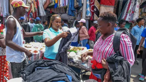 Anadolu via Getty Images A woman (R) in stripped pink shirt looks at trousers from a second-hand clothes vendor with long braids and in a blue T-shirt (C) at Gikomba market in Nairobi, Kenya. To the side of the vendor a man (L) in a vest and cap points - his side to the camera. Behind the women can be seen people walking past other stalls -September 2023.