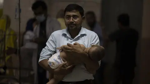 Getty Images A man wearing a button-up shirt and glasses stands in a hospital holding a baby in his arms