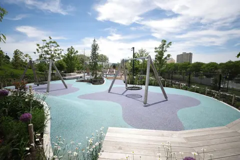 Talia Sprague/Bloomberg via Getty Images Two sets of swings are shown on a teal and purple playground surface. They are surrounded by lush landscaping and wooden benches and walking paths.