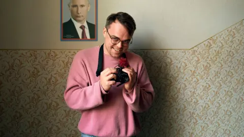 BBC/Made in Copenhagen/František Svatoš A man in a pink sweater holds a camera in front of a picture of Vladimir Putin