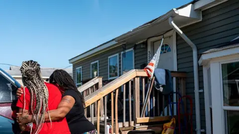 Getty Images Women hug near the entrance of a home. There's a US flag there. Some damage can be seen to the roof
