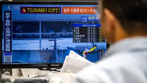Philip Fong / AFP via Getty Images The back of a person's head looks at a TV screen with the word 'tsunami' written and a live feed of a coastal area of northeastern Japan, in an office setting in Tokyo on Monday.