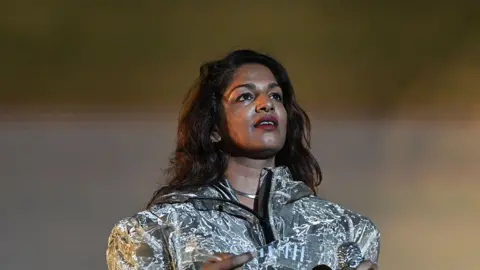 Getty Images The artist is wearing a shiny silver jacket. She has long wavy dark brown hair and brown eyes. She is wearing red lipstick and looking off into the top right corner of the camera.