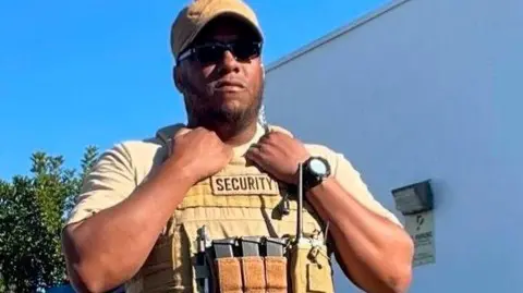 Islamic Center of San Diego Amin Abdullah stands outside wearing black sunglasses and a beige cap, T-shirt and tactical vest. 