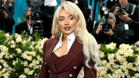 Getty Images Sabrina Carpenter wears a tailored maroon suit jacket with oversized lapel details and silver buttons, with blond waved hair over her shoulders - a bank of photographers are behind her 