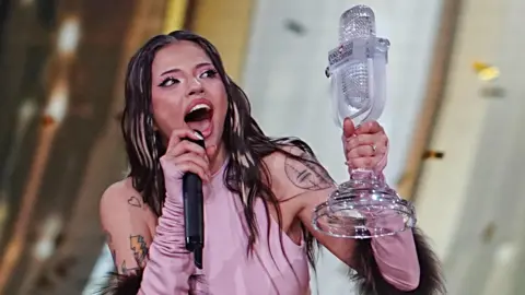 Getty Images Dara is holding Eurovision's glass microphone trophy after winning the competition in Vienna in May. She is wearing a pink top with her shoulders uncovered, showing several tattoos. She has blonde highlights in her otherwise dark hair and is open-mouthed with excitement. She has pink eyeshadow with dark eyeliner and thick eyelashes. She is talking into a black microphone.