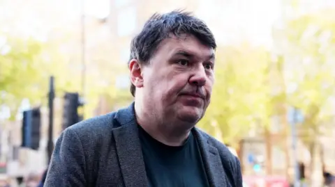 PA Graham Linehan, a man with dark hair, wearing a grey jacket and dark top.