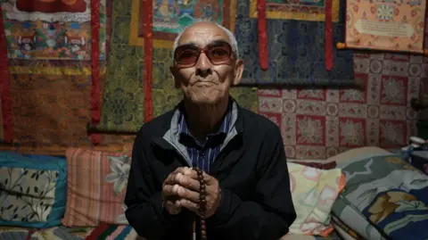 Sandeep Yadav/BBC Dawa Sangbo with sunglasses sits, wearing a black jacket over a blue striped shirt and holding prayer beads. The wall behind him is covered in colourful rugs. 
