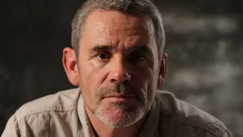 BBC/Arrow International Media/Steve Organ Paul Conroy poses for a headshot. He has short greying hair and stubble.