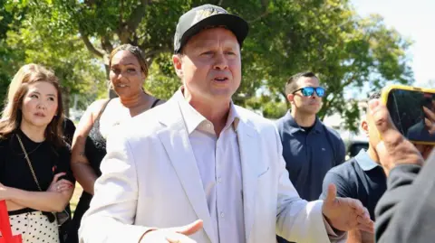 Getty Images Spencer Pratt speaks to voters on election campaign. He is wearing a black baseball cap and a white linen suit and white shirt. There is a crowd gathered around him to hear him speak.
