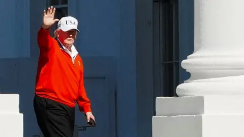 Reuters President Donald Trump, wearing a white USA cap and a red golfing sweater, waves as he passes by white pillars outside the White House