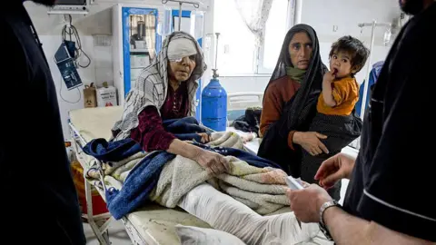 Getty Images Afghan woman with head and leg wrapped in bandage lies on a hospital bed flanked by another Afghan woman holding a baby
